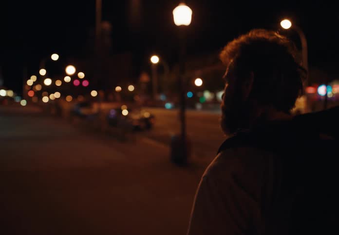 A man with a backpack walking the city streets at night with streetlights and vehicles