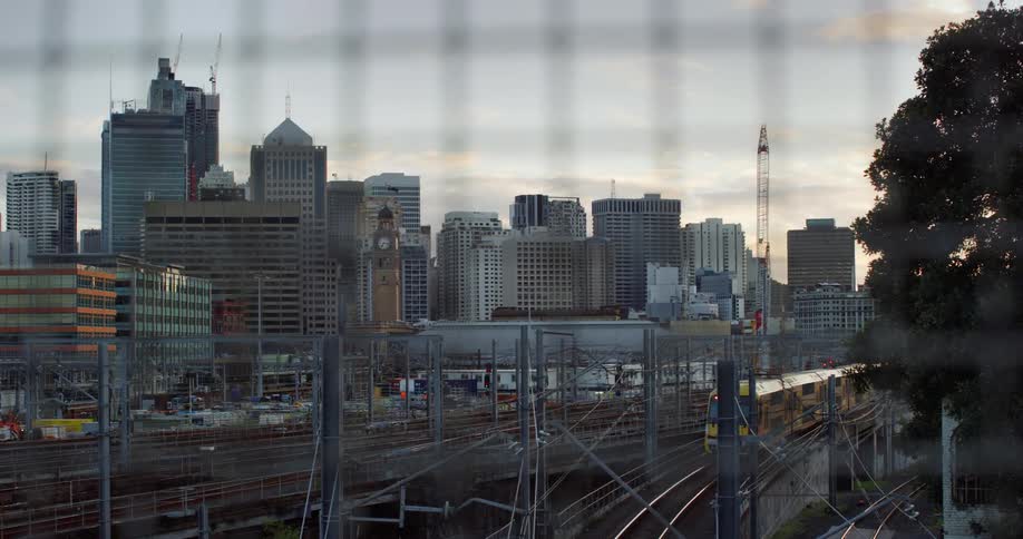 Train travels on the tracks past the trees as city buildings stand nearby