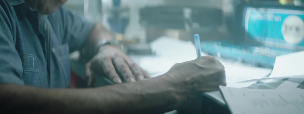 Man wearing a hat sits at a desk and writes on some paper