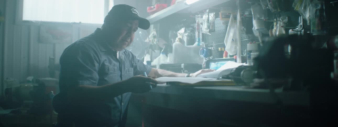 A mechanic sits inside a garage at a bench and writes on paperwork