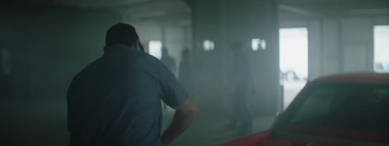 A mechanic works on a car in a garage as other mechanics walk past