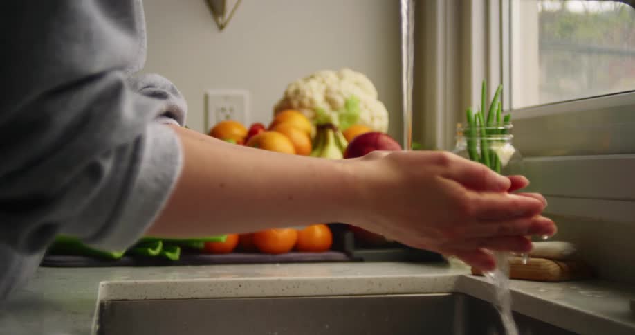 A person washes cherry tomatoes with soap in a kitchen faucet before adding it to a stack of produce