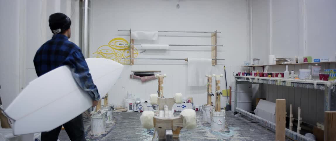 Man carries a white surfboard across the workshop