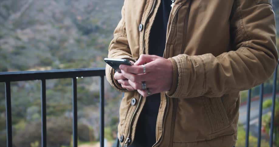 A white man in a coat stands outdoors on a balcony scrolling through a smartphone