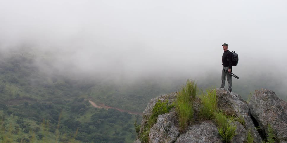 A man, who is holding a tripod, standing on top of boulders on a foggy day