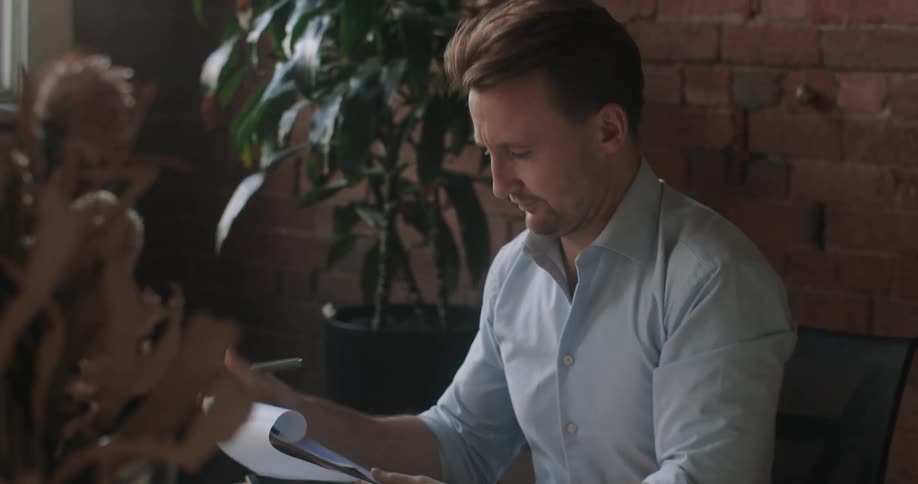 A man sitting at his desk in his office talks to someone on his computer while looking at paperwork
