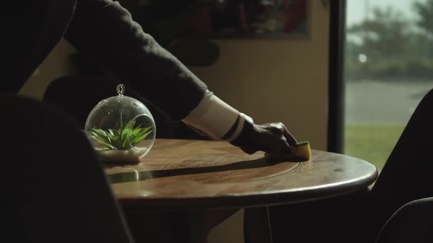 A person with a spray cleaner and a sponge wiping down a table surrounded by chairs