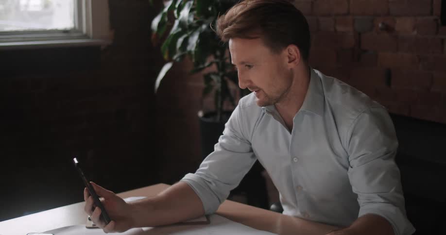 A man sits at a table with a large paper spread across it while video chatting on a smartphone