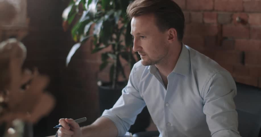 A man sitting at a desk talks during a video meeting on his laptop and takes notes on a piece of paper