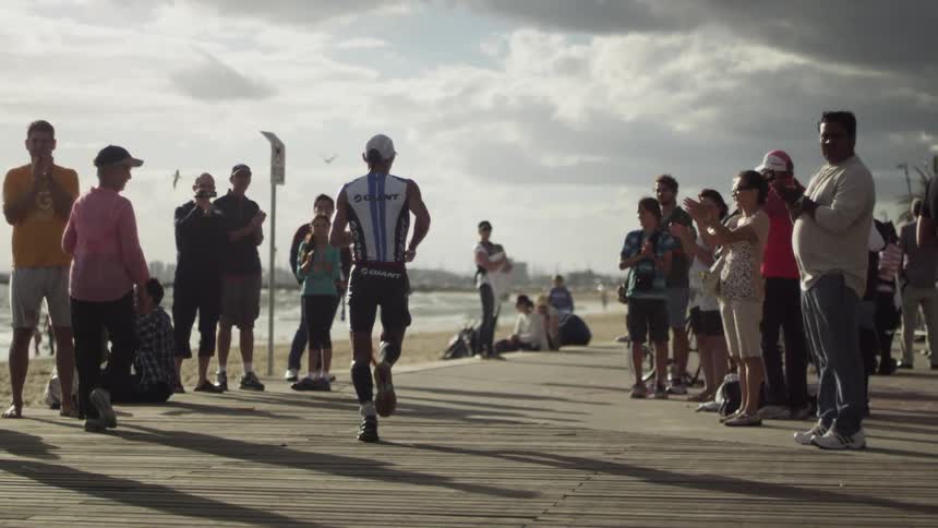 Men running a marathon while people stand and clap with the beach and ocean in the background