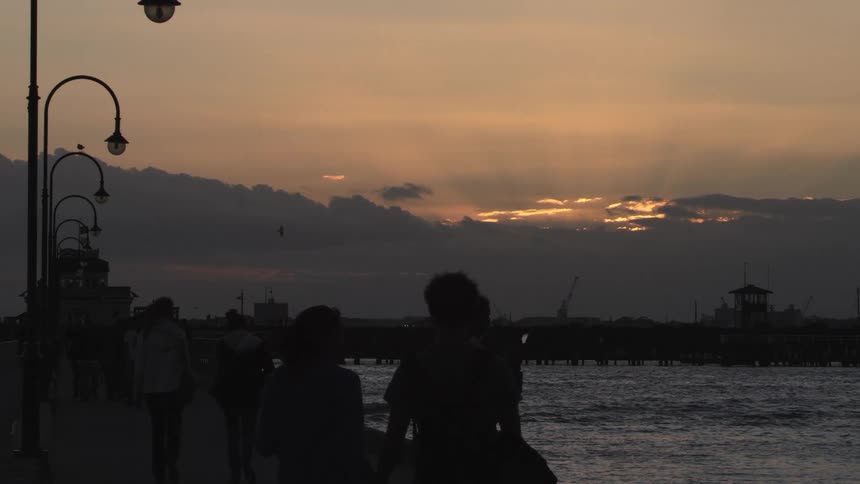 People and couples walk along the waterfront holding hands as birds fly over the water at sunset