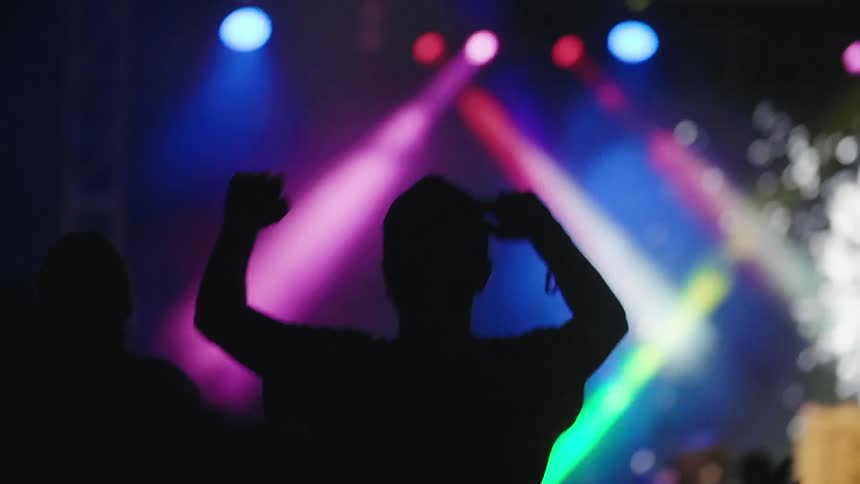 A concert crowd in silhouette with arms and hands waving in the air as a laser light show happens on stage