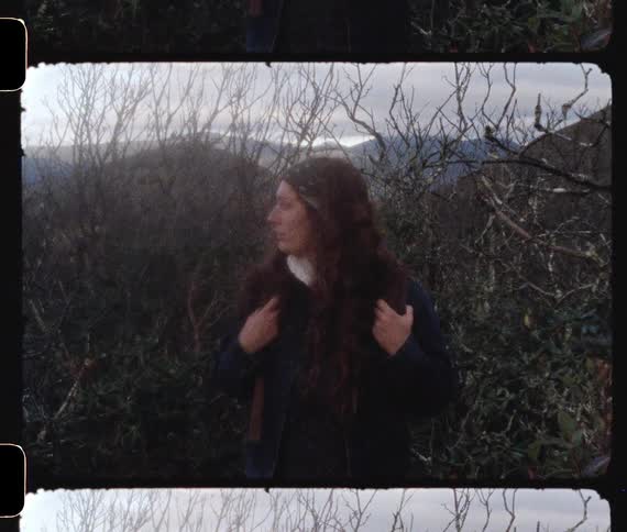 A woman stands in front of some scrub brush near some mountains