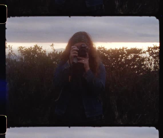 Woman holds a camera up to her face to take a photograph while standing in front of some trees
