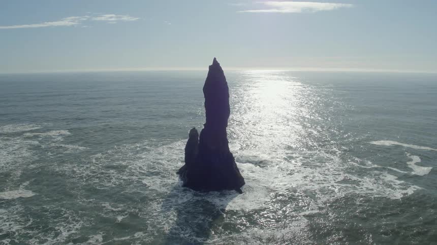 Ocean waves swirl and splash against rock formations near the coast as sunshine shimmers on the surface