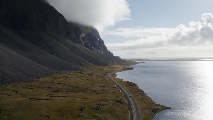 Cars follow road bordering lake near towering mountain topped by clouds