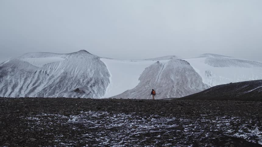 A person with a backpack walking on barren terrain with a snow covered mountain and the sky in the background