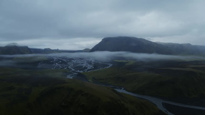 Low clouds hover over hills with streams running between as a mountain looms in the distance
