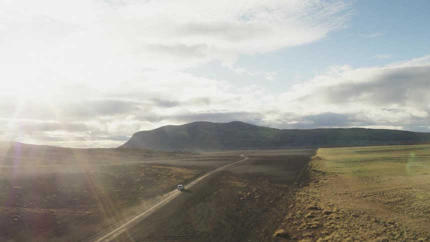 A vehicle driving on a dirt road with a mountain range the sky and clouds in the background