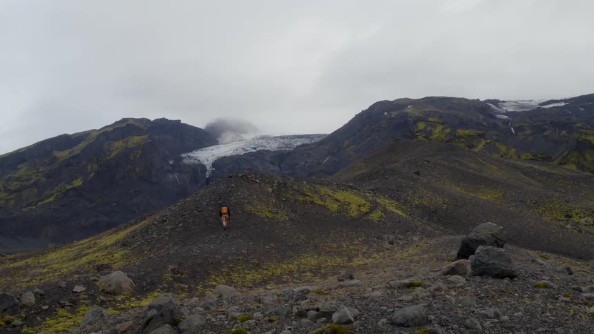 A man walking up a hill with mountains the sky and clouds in the background