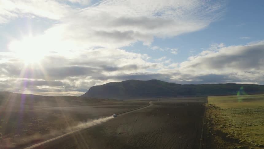 A car traveling on a dirt road leaves a dust trail as it heads toward mountains on a sunny day