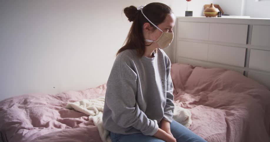 Woman sits on bed wearing face mask looking toward the window