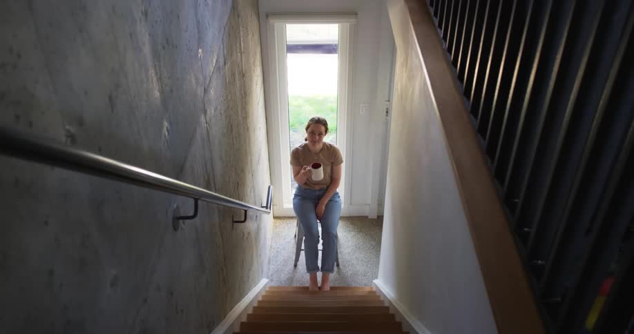 A woman is drinking from a mug at the bottom of the stairs