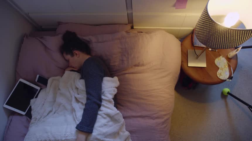 Birds eye view of a young woman lying in bed with her ipad and phone and then peeking under the sheet