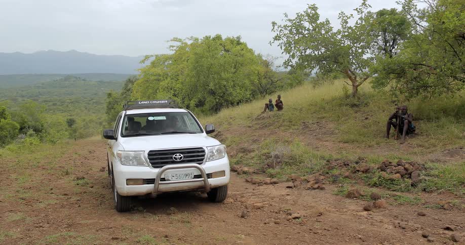 A vehicle is driving up a dirt road while villagers sit on the sides watching