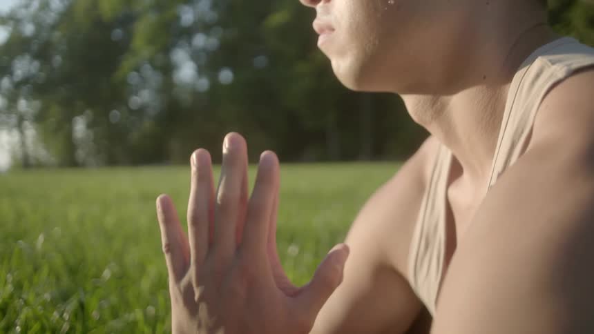 An asian man sitting in a yoga posture outdoors in a field on a sunny day