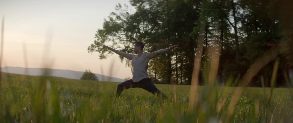 A man stretching in a grassy field with trees and the sky in the background