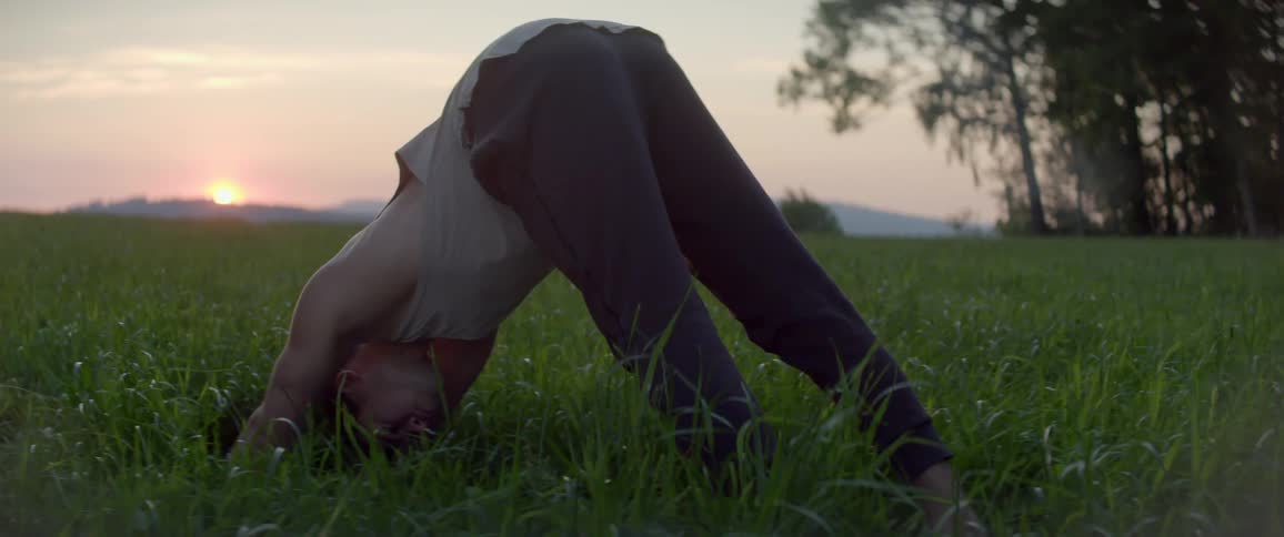 A man stretching on a grassy field with the sky the sun and clouds in the background