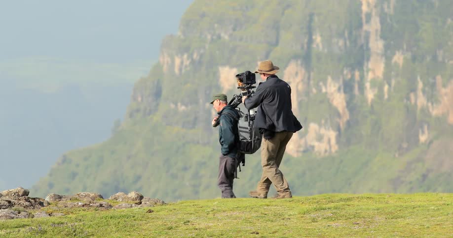 Two men carrying cameras are walking over a hill