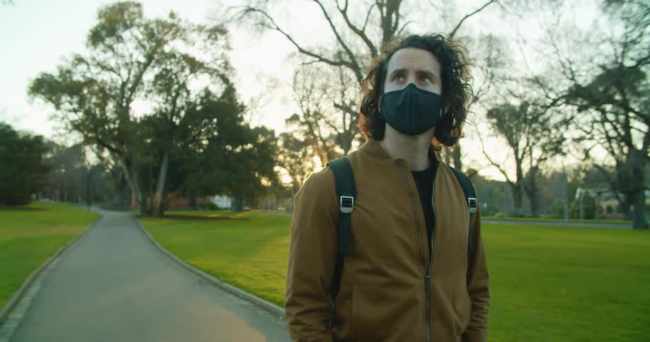 A man in a black medical mask stands on a walkway in a park looking around at the surroundings