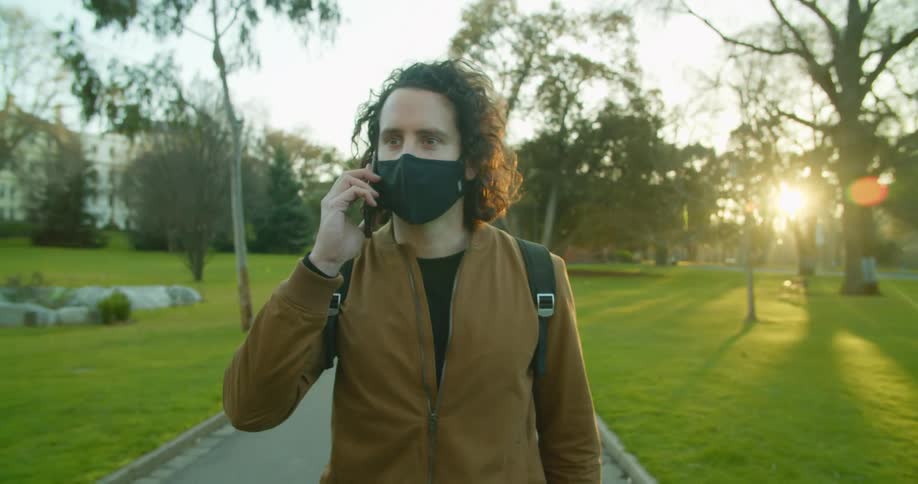 A man wearing a mask talks on his phone as he walks on a path through a park