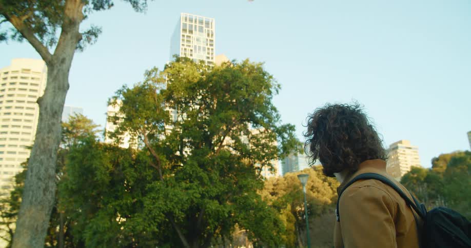 A man in black medical mask and brown jacket stands in a green space in a city park looking leisurely at a tree