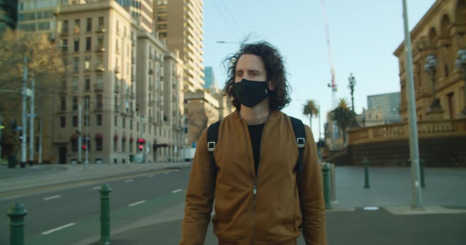 A man in a black mask walks down the sidewalk next to a wide street in a metropolis during the day