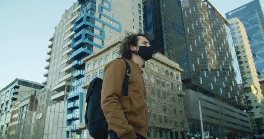 A man stands on a corner in a downtown area of a metropolis looking up at a skyscraper before turning to gaze around the area