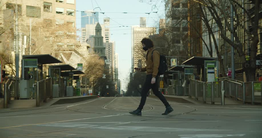 Man wearing face mask crosses empty street near downtown taking mobile phone out
