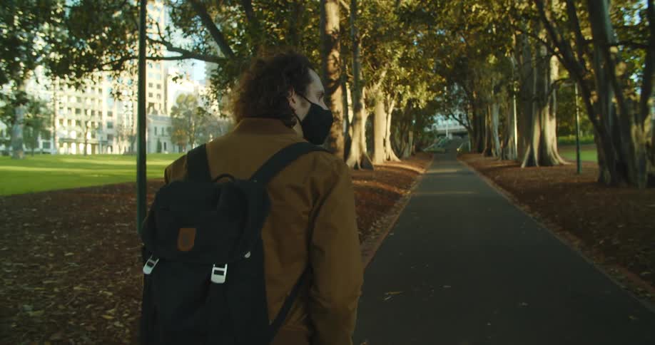 A man with a backpack and a mask walking in a pathway with buildings in the background
