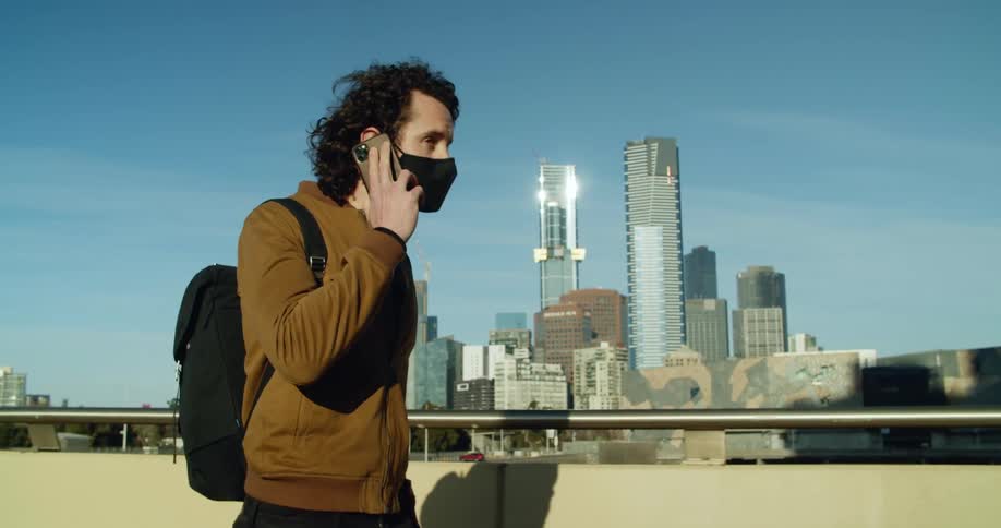 A man with a mask talking on a smartphone with buildings and skyscrapers in the background