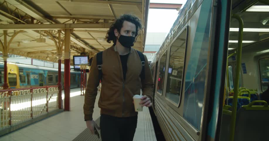 A man wearing a mask and carrying a cup of coffee checks his phone before getting on a train