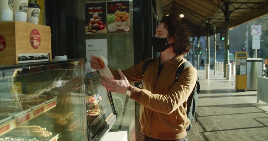 A man wearing a black medical mask recieves food and drink from a food stand before walking off down the sidewalk