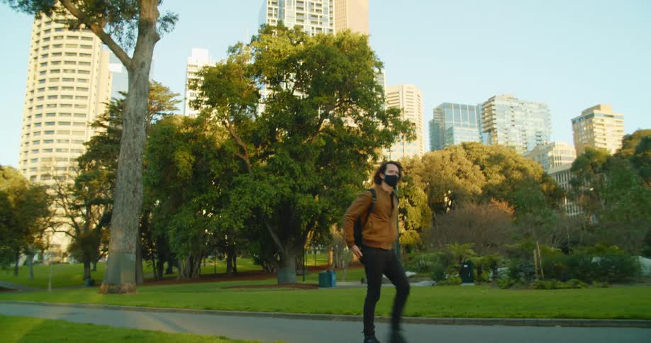 A man with a mask walking in a park and looking at a smartphone with buildings and skyscrapers in the background