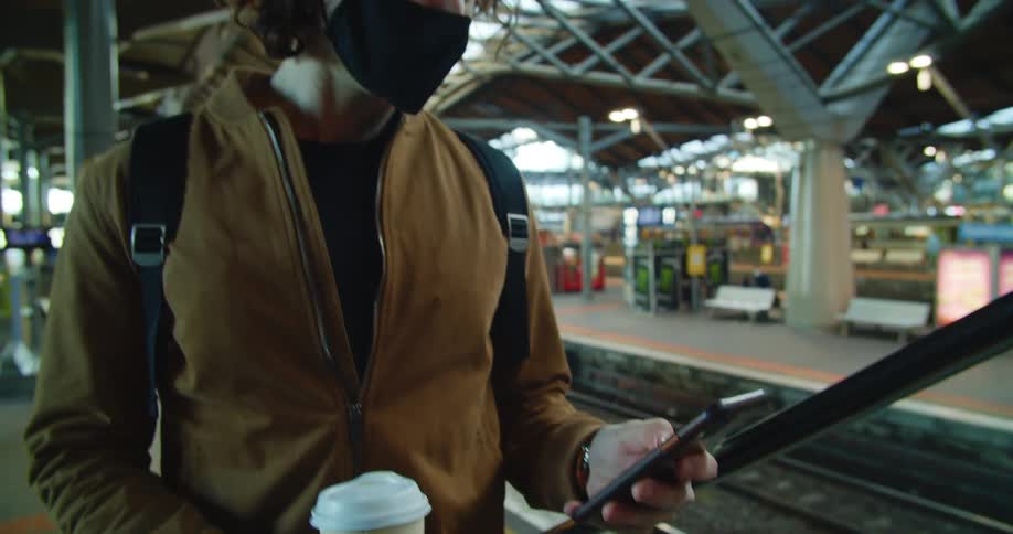Wearing a face mask holding a to go coffee cup and texting on mobile phone man waits near tracks in transit station