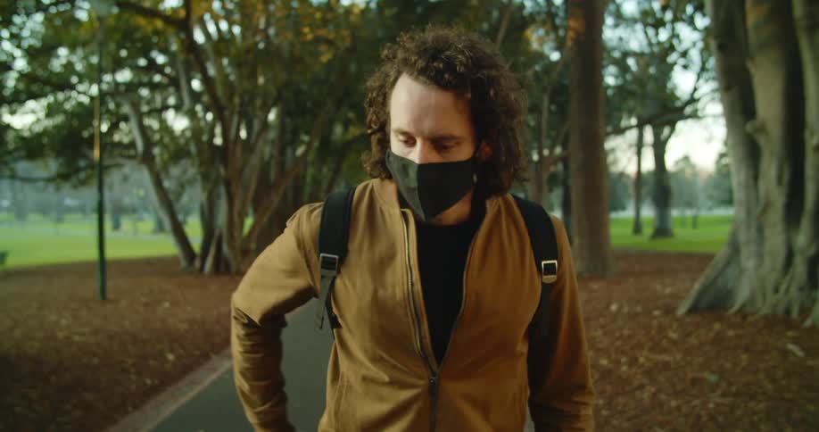 A man wearing a mask and walking on a path in the park pulls his phone out of his pocket and looks at it