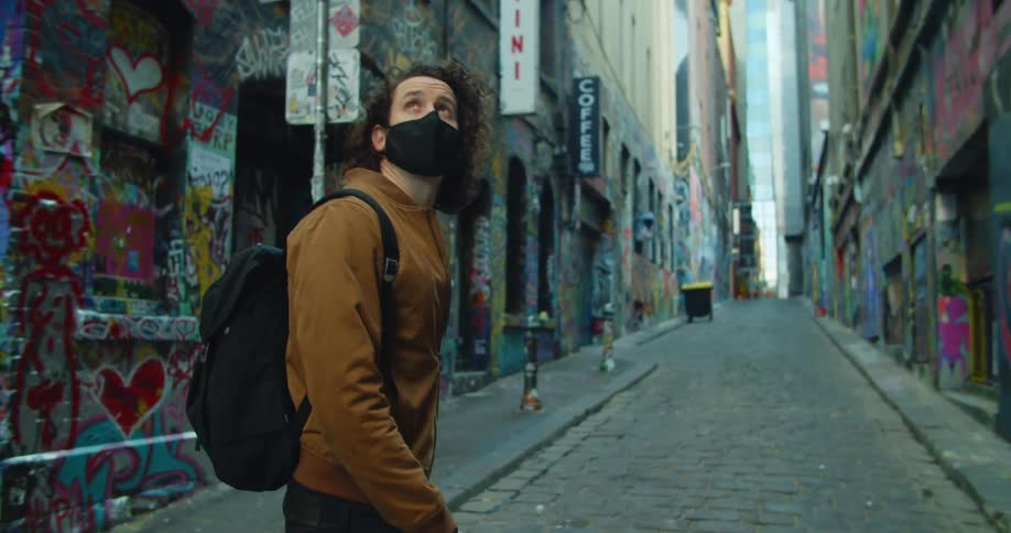A man wearing a black medical mask walks down an alley covered in colorful graffiti while turning his head to look up on the buildings on either side