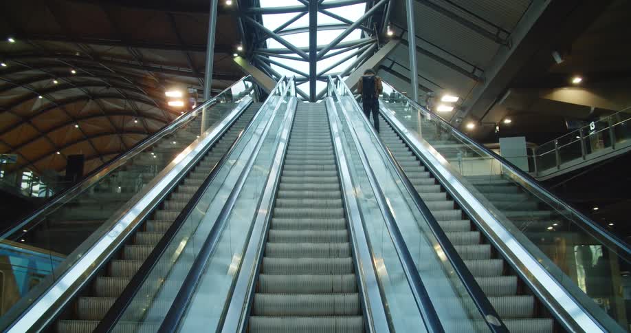 A man rides an outdoor escalator as a train passes by under an atrium