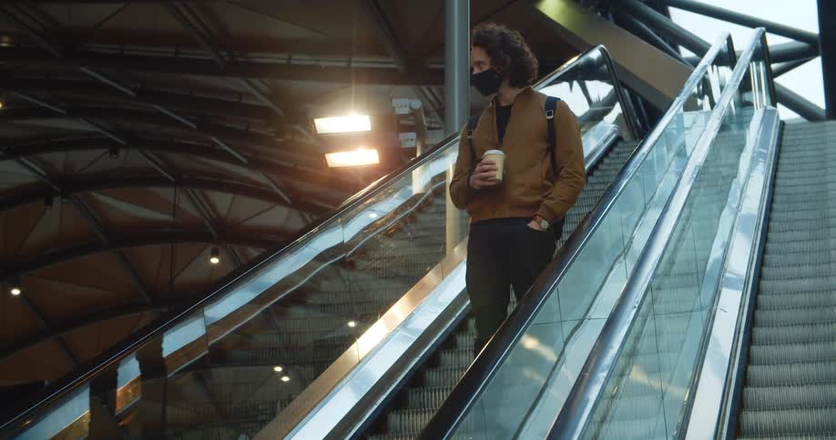 A man carrying a coffee cup and wearing a mask rides down an escalator