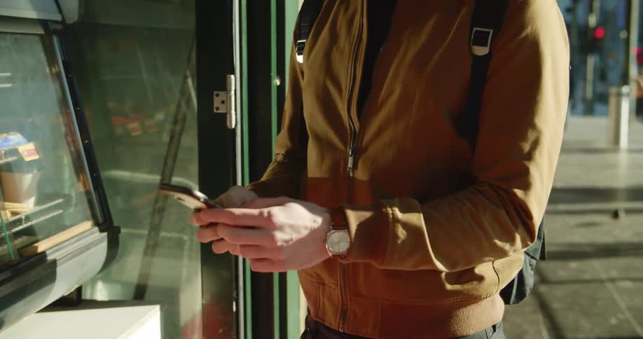A man wearing a mask standing in front of a store on a city sidewalk takes out his phone and texts